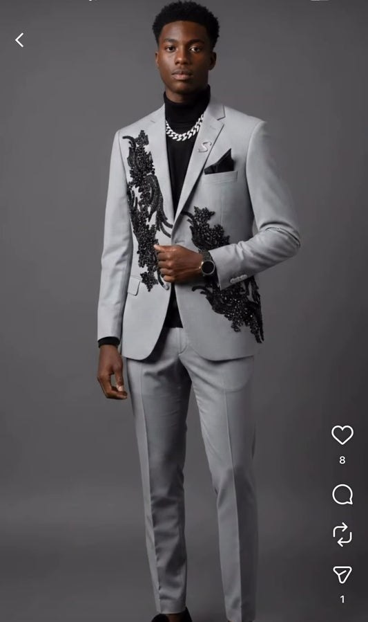 A man poses against a gray background in the Unique Design Menswear custom prom suit—grey with black rhinestones—styled with a black shirt, chain necklace, and wristwatch.