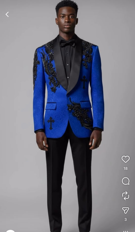 A man models the Unique Design Menswear custom prom suit—a royal tuxedo with black rhinestones—featuring black lapels, cross designs, and bead embellishments, paired with black pants and a bow tie against a gray background.
