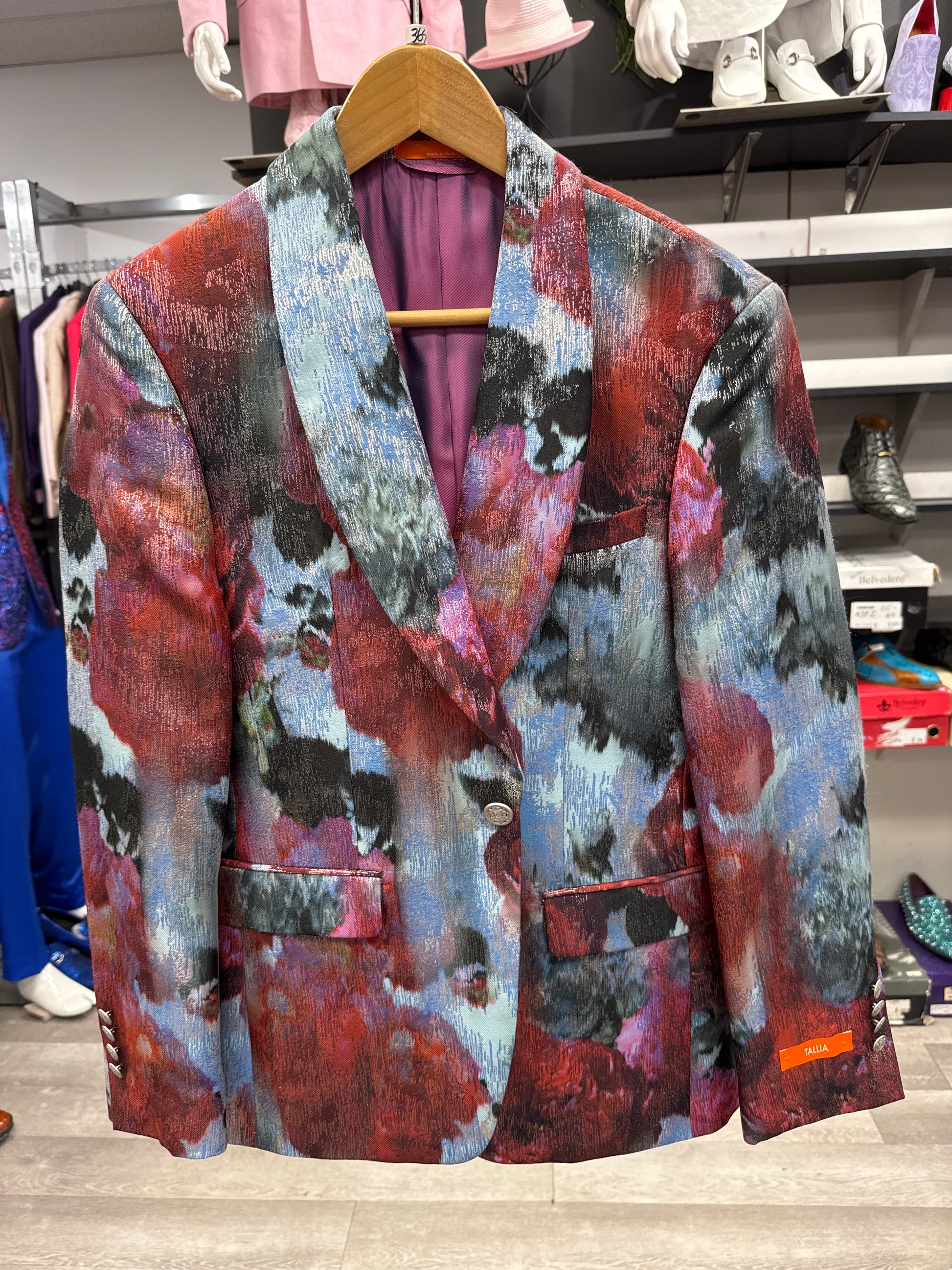 The EJ SAMUEL JACKET 18 CLEARANCE, featuring a vibrant abstract pattern in red, blue, black, and gray, is displayed on a wooden hanger in the store. Please note: this item is final sale—no returns or exchanges will be accepted.