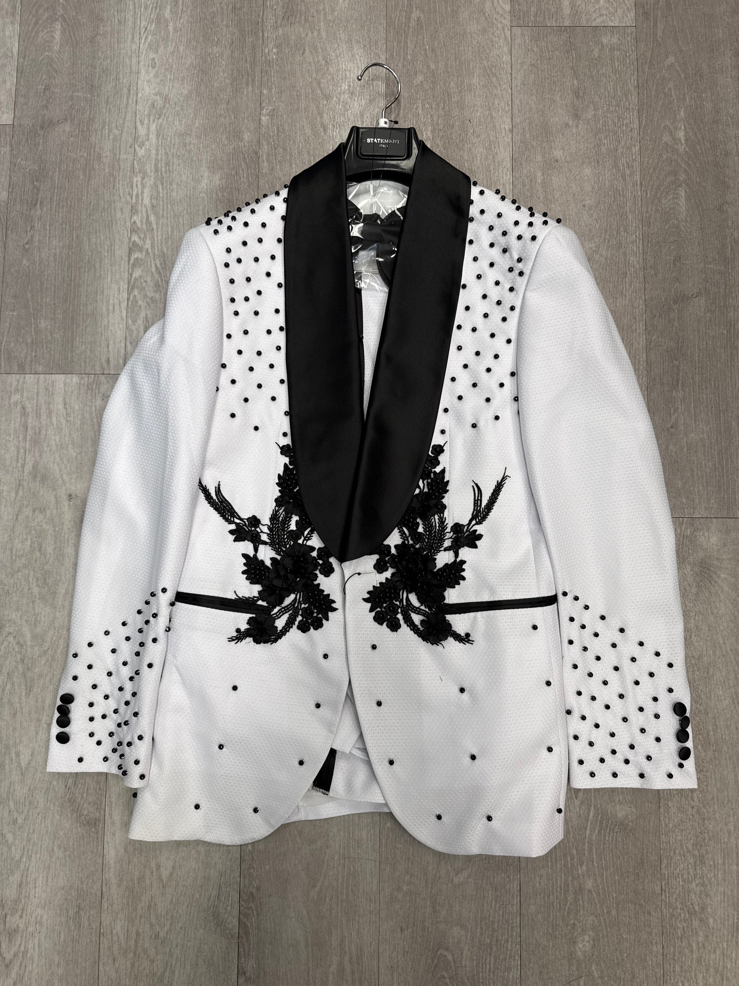 The STATEMENT CLOTHING | ELIO-WHITE WITH BLACK STONES tuxedo blazer by Statement Clothing features black floral embroidery, black lapels, and scattered black studs, displayed on a hanger against a wooden floor.