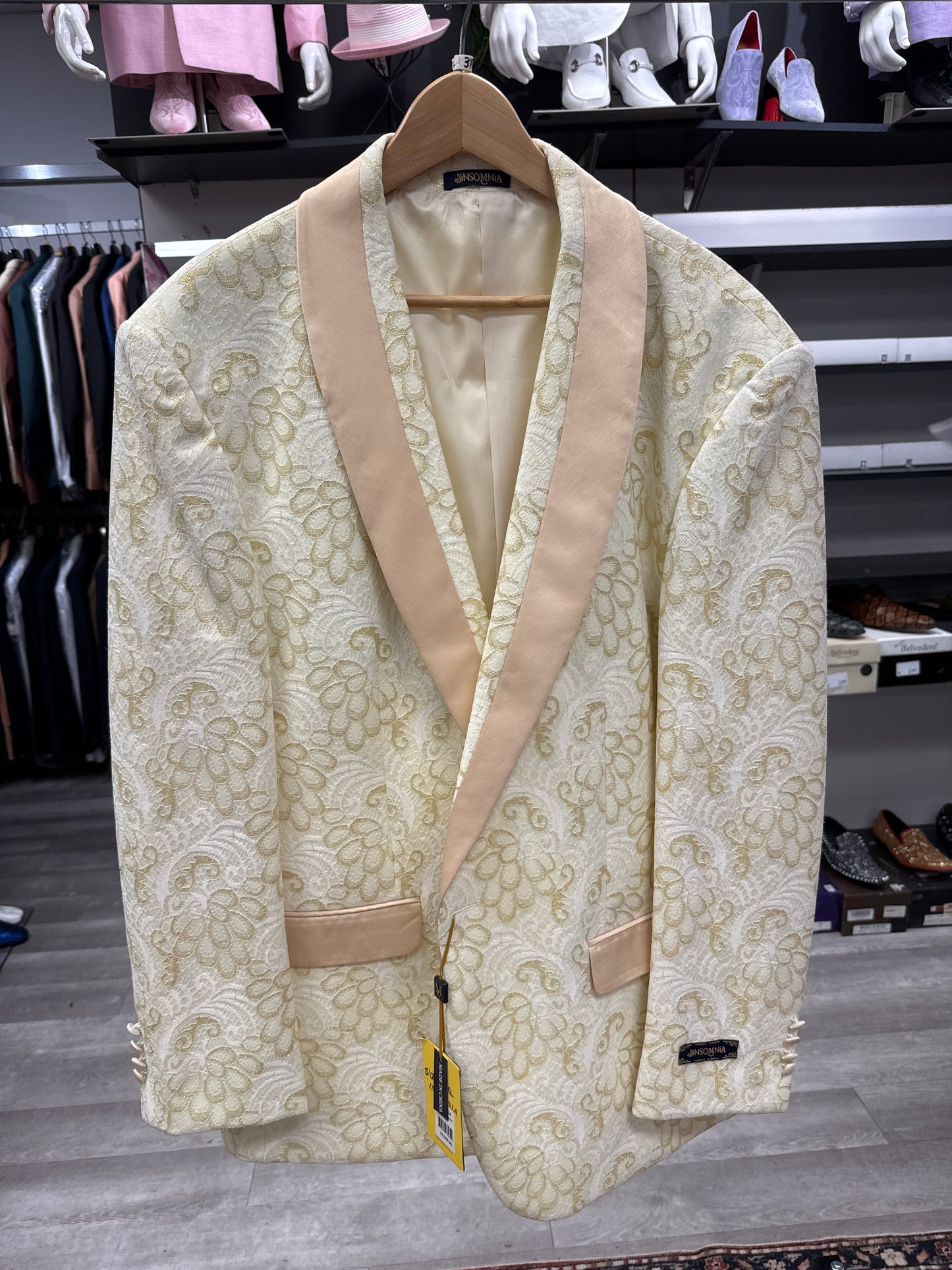 A cream-colored men’s blazer—JACKET 28 CLEARANCE by EJ SAMUEL—features a floral pattern and beige satin lapels, and is displayed on a wooden hanger in a clothing store. Please note, this item is final sale: no returns or exchanges accepted.