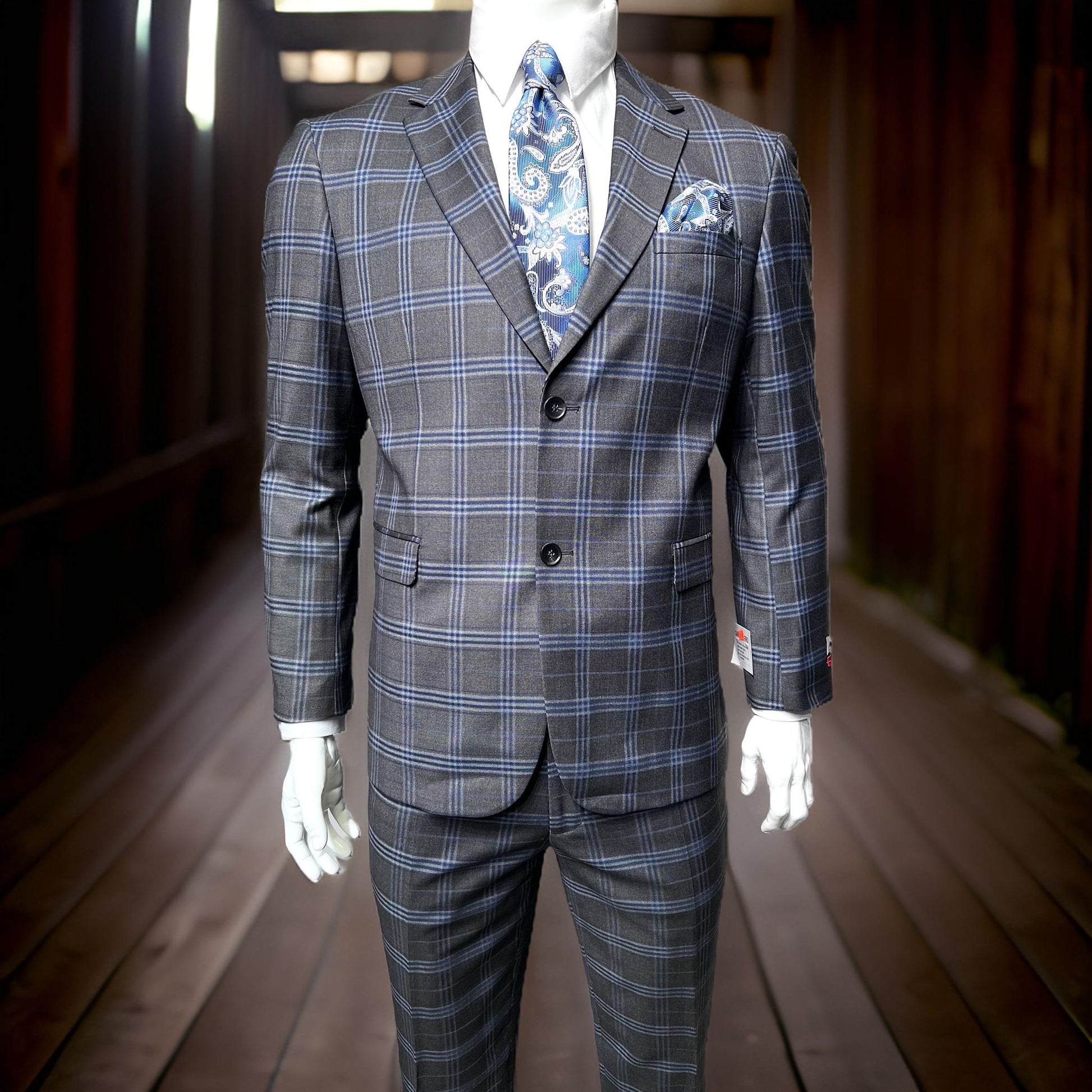 A mannequin showcases the Unique Design Menswear Gianco Ferro Modern Fit Suit Grey 7102/17 (52R ONLY) with a blue patterned tie and pocket square, standing on a wooden walkway in low lighting.
