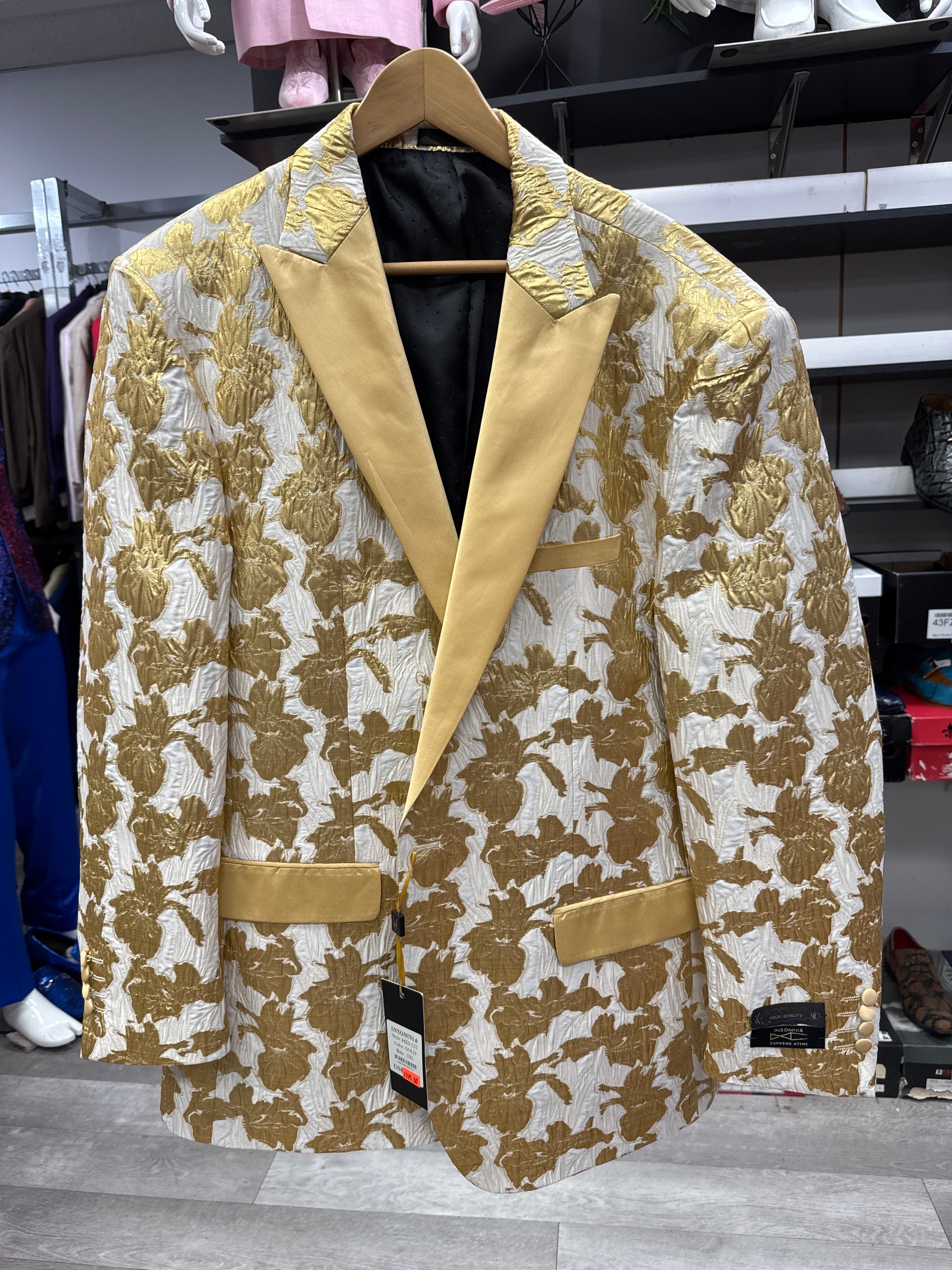The EJ SAMUEL JACKET 4 CLEARANCE, featuring a gold and cream floral pattern with solid gold lapels and pockets, hangs on a wooden hanger in a clothing store. Please note: this item is final sale—no exchanges or returns permitted.