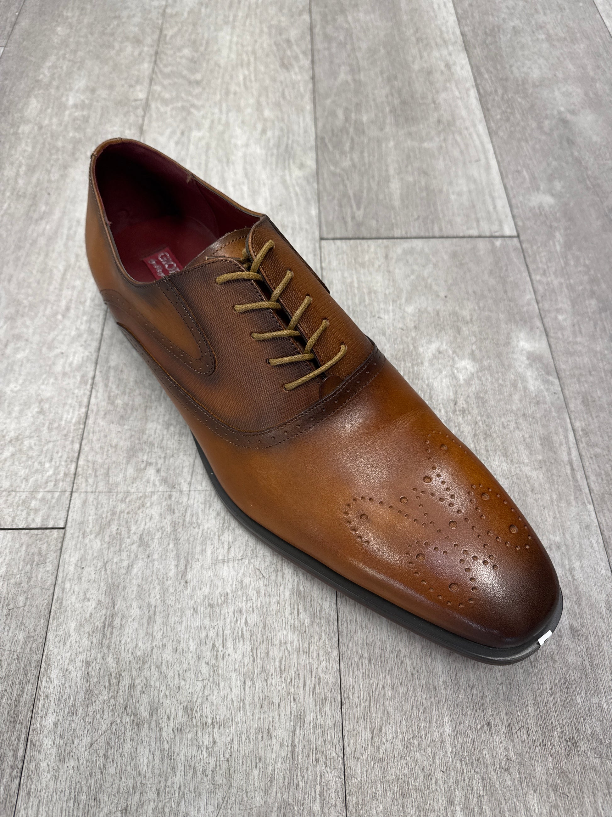 A Fiesso brown leather Shoe 92 in size 15, featuring decorative perforations on the toe, is elegantly displayed on a light wood floor. Please note, this item is FINAL SALE and cannot be returned.