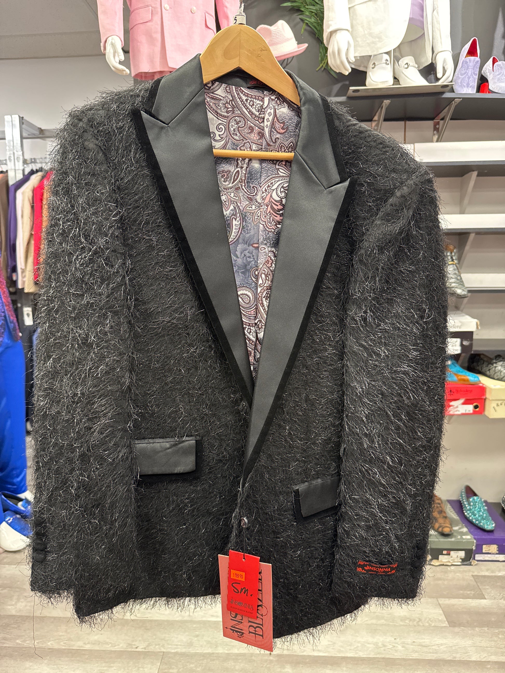 An EJ SAMUEL JACKET 12 CLEARANCE—featuring a black fuzzy finish, satin lapels, and paisley lining—hangs on a wooden hanger in a clothing store; please note, this item is final sale.