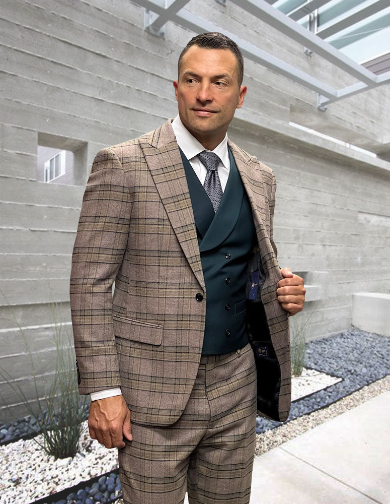 A man wearing the STATEMENT CLOTHING | AMICCI-JADE brown plaid suit with peak lapel and dark green vest and tie from Statement Clothing stands in a modern outdoor setting with concrete walls and decorative stones.