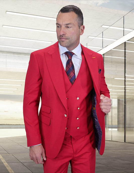 A man in a modern-fit STATEMENT CLOTHING | AMIRI-BERRY red wool suit by Statement Clothing, paired with a white shirt and red plaid tie, stands inside a sleek concrete building with large windows.