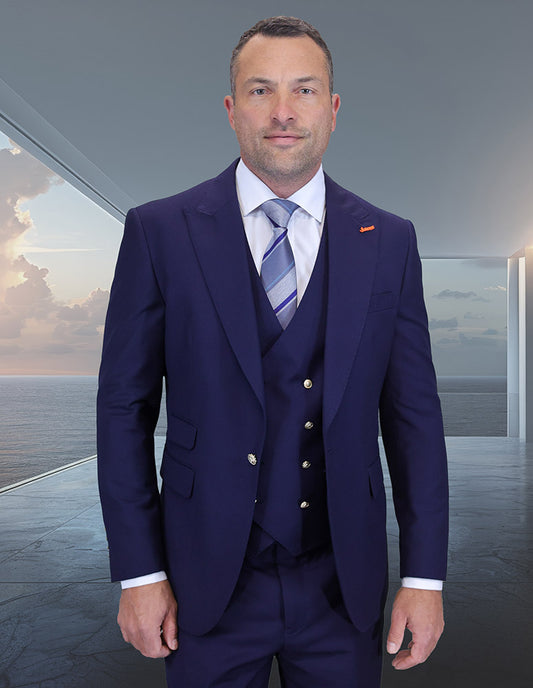 A man in the modern fit STATEMENT CLOTHING | AMIRI-NAVY Italian wool suit by Statement Clothing, paired with a striped tie and white shirt, stands indoors near large windows overlooking the ocean at sunset.