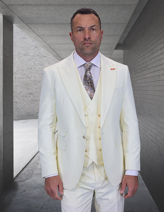 A man in the STATEMENT CLOTHING | AMIRI-OFFWHITE suit by Statement Clothing, featuring a modern fit, cream vest, and patterned tie, stands in a concrete hallway with gray brick walls, looking directly at the camera.