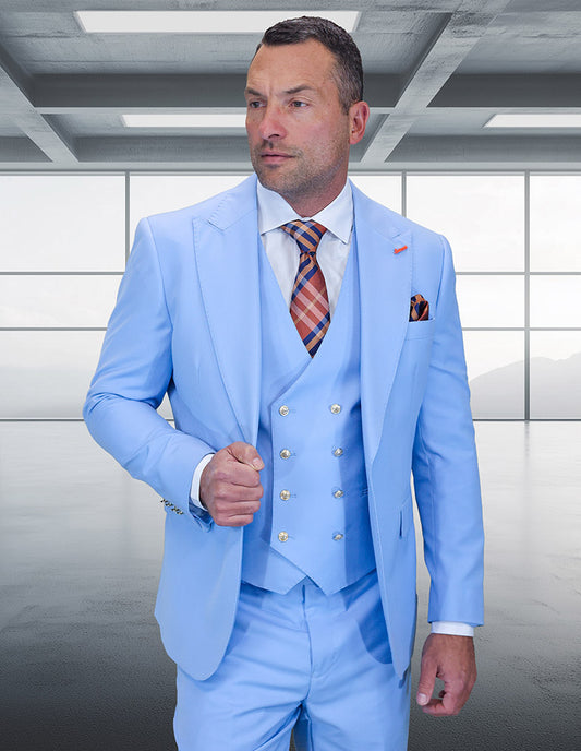 A man wearing the STATEMENT CLOTHING | AMIRI-SKY BLUE suit by Statement Clothing, paired with a white shirt and plaid tie, stands in a modern empty room with large windows.