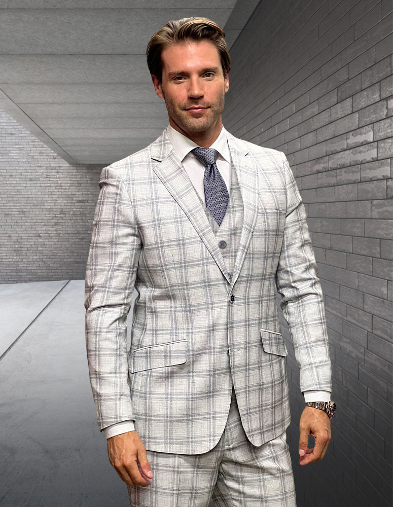 A man wearing the STATEMENT CLOTHING | ANGEL-1-NATURAL modern fit Italian wool suit by Statement Clothing with flat front pants, vest, and tie stands in a sleek gray-brick hallway, looking at the camera.
