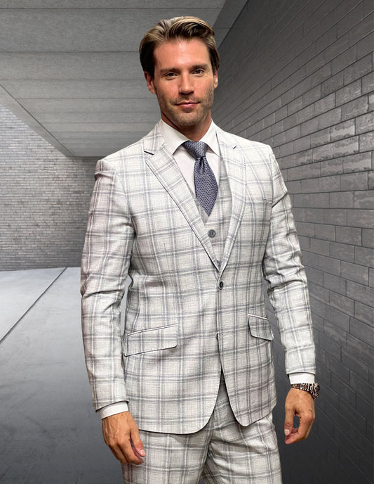 A man wearing the STATEMENT CLOTHING | ANGEL-1-NATURAL modern fit Italian wool suit by Statement Clothing with flat front pants, vest, and tie stands in a sleek gray-brick hallway, looking at the camera.