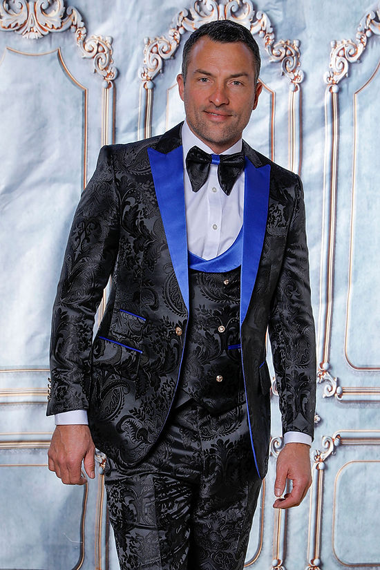 A man in a Manzini Armani 3-Piece Slim Fit Satin Filigree Pattern Black/Royal Tuxedo Prom Suit stands before an ornate, light-colored backdrop, wearing a white shirt and bow tie.