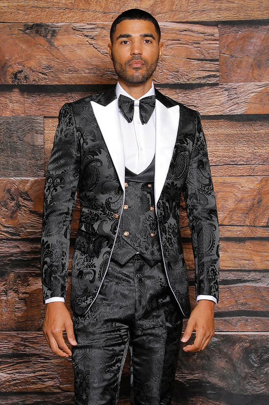 A man wears a Manzini Armani 3-Piece Slim Fit Satin Filigree Pattern Black/White Tuxedo Prom Suit with a white dress shirt and black bow tie, standing before a wooden background.