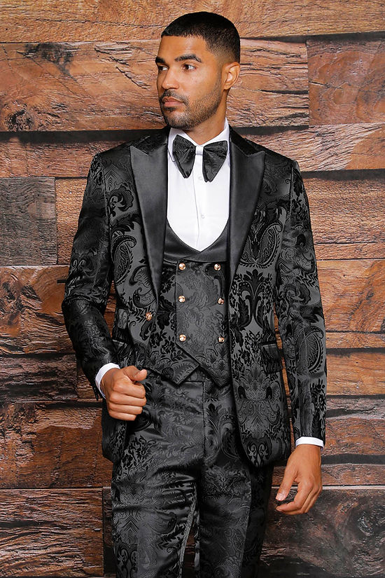 A man wears the Manzini Armani 3-Piece Slim Fit Satin Filigree Pattern Black Tuxedo Prom Suit, featuring a white shirt, black bow tie, and waistcoat, standing against a wooden wall.