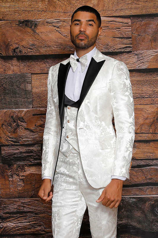A man wears the MANZINI ARMANI 3-Piece Slim Fit Satin Filigree Pattern Cream/Black Tuxedo Prom Suit by Manzini, paired with a white shirt and bow tie, standing before a wood-textured background.