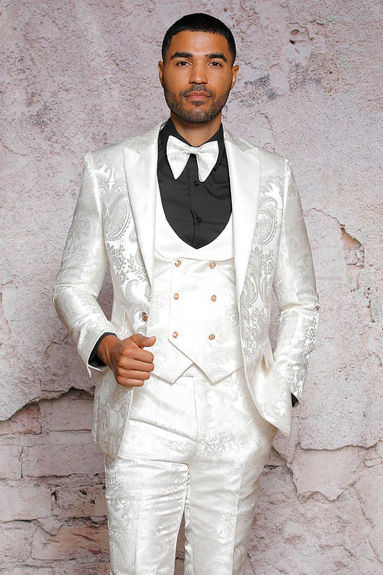 Man wearing the Manzini Armani 3-Piece Slim Fit Satin Filigree Pattern Cream Tuxedo Prom Suit by Manzini, with matching vest, black shirt, and white bow tie, standing against a textured light stone wall.