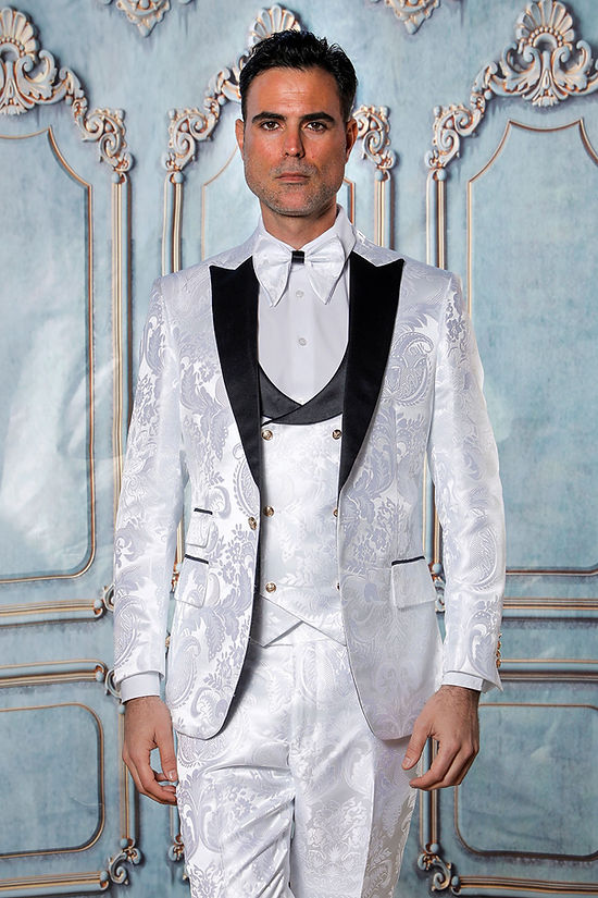 A man wears the Manzini Armani 3-Piece Slim Fit Satin Filigree Pattern White/Black Tuxedo Prom Suit, standing before an ornate light blue and gold wall.