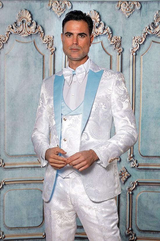 A man wears the Manzini Armani 3-Piece Slim Fit Satin Filigree Pattern White/Sky Tuxedo Prom Suit, featuring light blue lapels and a floral vest, standing before an ornate decorative wall.