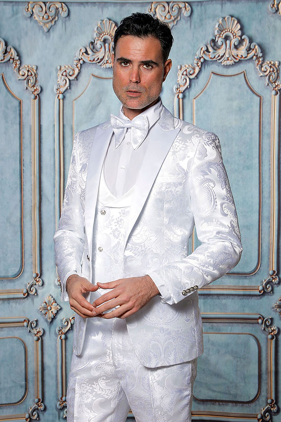 A man models the Manzini Armani 3-Piece Slim Fit Satin Filigree Pattern White/White Tuxedo Prom Suit by Manzini, posing in front of an ornate pale blue and gold backdrop.