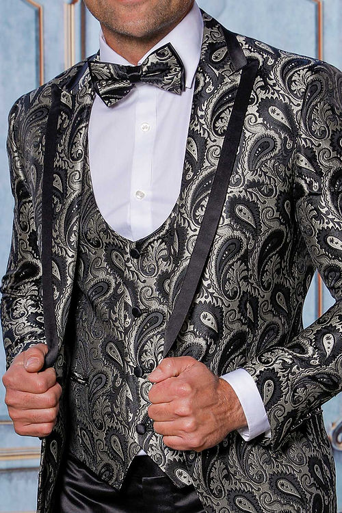 A man in an Insomnia MANZINI ASHER 3-piece slim fit shiny satin paisley black tuxedo with a shawl lapel and bow tie stands before an ornate backdrop.
