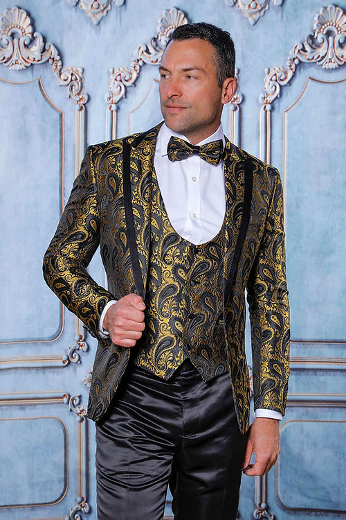 A man models the Insomnia MANZINI ASHER 3-Piece Slim Fit Shiny Satin Paisley Gold Tuxedo Prom Suit with a matching bow tie, standing against an ornate blue backdrop to highlight its intricate design.