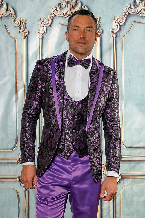 A man in an Insomnia MANZINI ASHER 3-piece slim fit shiny satin paisley purple suit, featuring a peak lapel jacket and matching vest with a bow tie, stands before an ornate backdrop.