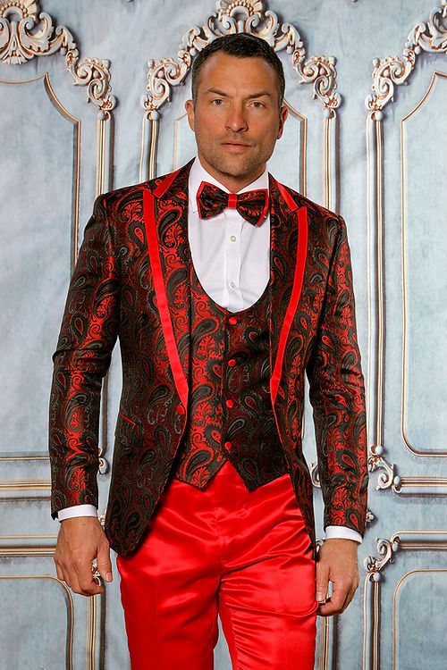 A man poses against an ornate backdrop in the Insomnia MANZINI ASHER 3-Piece Slim Fit Shiny Satin Paisley Red Tuxedo Prom Suit, accompanied by a matching bow tie and red trousers.