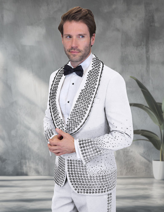 A man in the STATEMENT CLOTHING | AWARD-WHITE tuxedo by Statement Clothing, paired with a black bow tie and white shirt, stands before a gray textured background next to a potted plant.