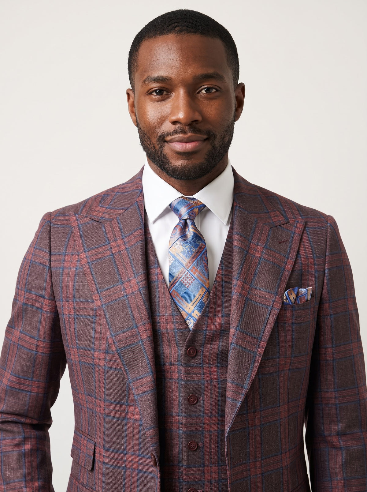 A man wears a Tiglio Luxe Antibes Brick/Blue Plaid Semi Wide Leg Suit TL8305 featuring a brick-red and blue plaid three-piece design, white dress shirt, patterned blue tie, and matching pocket square.