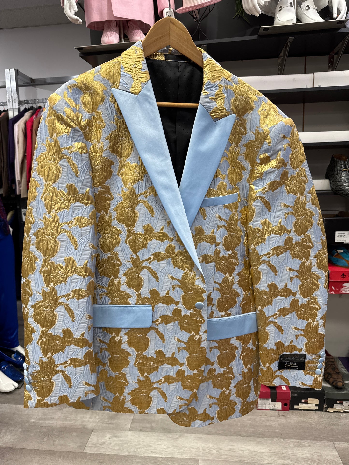 The EJ SAMUEL JACKET 5 CLEARANCE, a gold floral-patterned blazer featuring light blue lapels and pocket accents, hangs on a wooden hanger in the clothing store; please note this is a final sale item with no exchanges or returns.