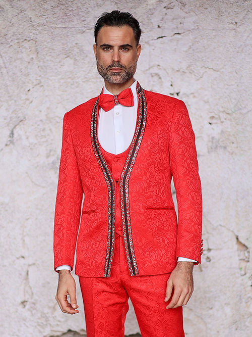 A man confidently stands against a textured wall in an Insomnia MANZINI BEGGIO 3-Piece Slim Fit Glitter Trim Red Tuxedo Suit, with a white shirt and matching bow tie.