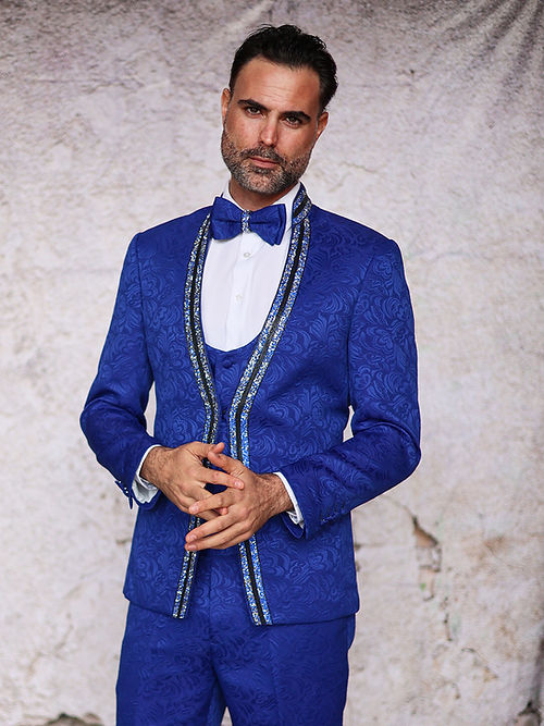 A man in a MANZINI BEGGIO 3-Piece Slim Fit Glitter Trim Royal Tuxedo Suit by Insomnia, paired with a matching bow tie and subtle floral pattern, poses against a textured backdrop.