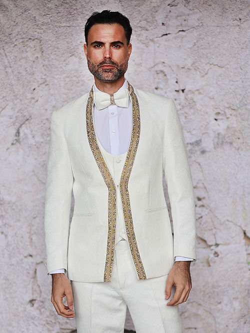 A man wearing an Insomnia MANZINI BEGGIO 3-Piece Slim Fit Glitter Trim White Tuxedo Suit stands against a textured stone wall.