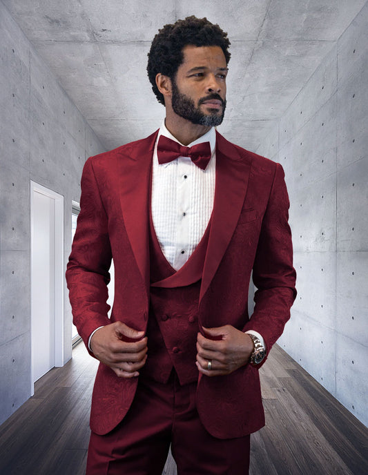 A man with short curly hair and a beard wears the Statement Clothing BELAGIO15-BURGUNDY suit with flat front pants, a burgundy bow tie, and a watch, standing in a modern hallway with concrete walls and wooden floor.