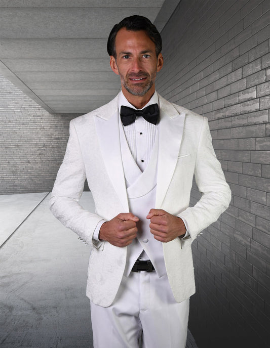 A man stands indoors wearing the STATEMENT CLOTHING | BELAGIO15-WHITE tailored fit tuxedo with flat front pants by Statement Clothing, paired with a black bow tie. He holds his jacket front against a backdrop of gray brick and concrete walls.