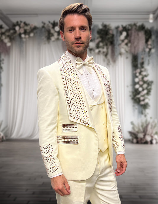 A man wearing the STATEMENT CLOTHING | BELLA-OFFWHITE tuxedo by Statement Clothing, featuring rhinestone details and subtle burgundy accents, stands in an elegant indoor space with white drapery and floral decor.