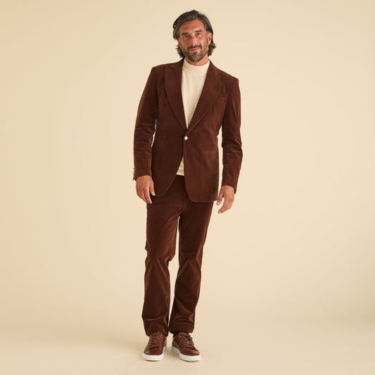 A man stands against a beige background wearing the Inserch Velvet Suit BL007-131 Swiss Chocolate by Inserch, paired with a cream turtleneck sweater, modern fit pants, and brown sneakers.