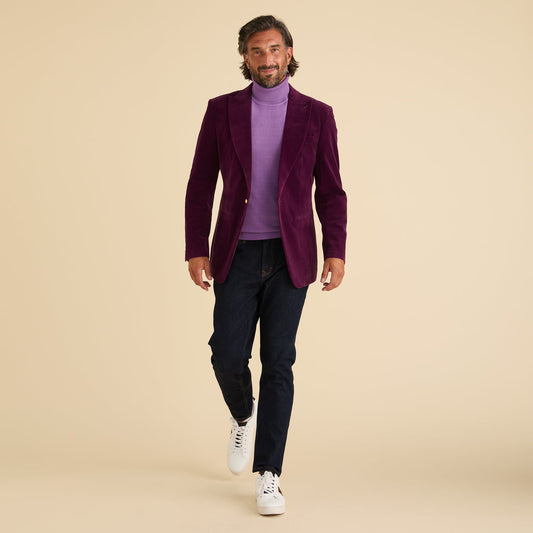 A man in the Inserch Velvet Blazer BL007-166 Deep Violet with a peak lapel, lavender turtleneck, dark jeans, and white sneakers walks toward the camera against a plain beige background.