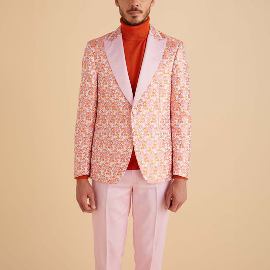 A man in the Inserch Floral Jacquard Blazer BL203-62 Pink by Inserch, paired with light pink pants and an orange turtleneck, stands against a beige background.