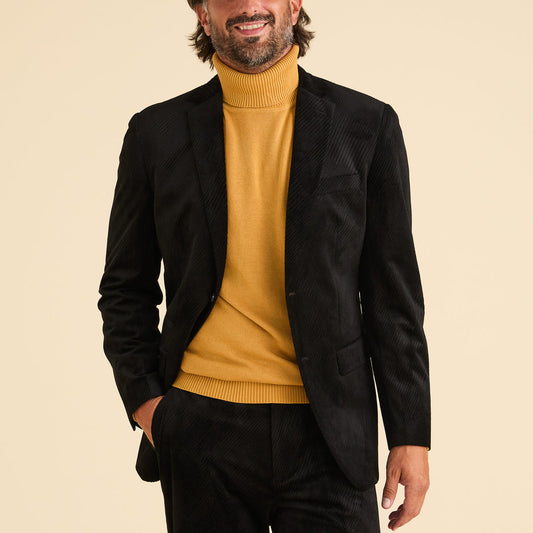A man wearing the Inserch Velvet Diagonal Blazer BL275-01 Black with matching pants and a mustard yellow turtleneck, stands against a plain beige background.