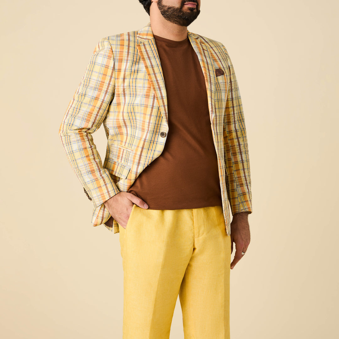 A man wearing the Inserch Seersucker Check Blazer BL277-00055 Yuzu by Inserch, paired with a brown shirt and yellow pants, stands against a beige background.