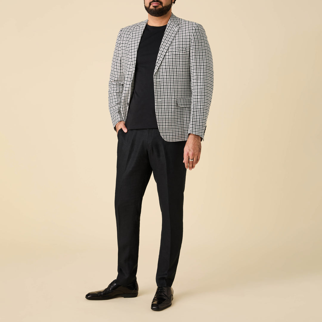 A man wearing the Inserch Houndstooth Linen Blazer BL279-00041 Black & White by Inserch, along with black t-shirt, pants, and shoes, stands against a beige background. His face is not visible.