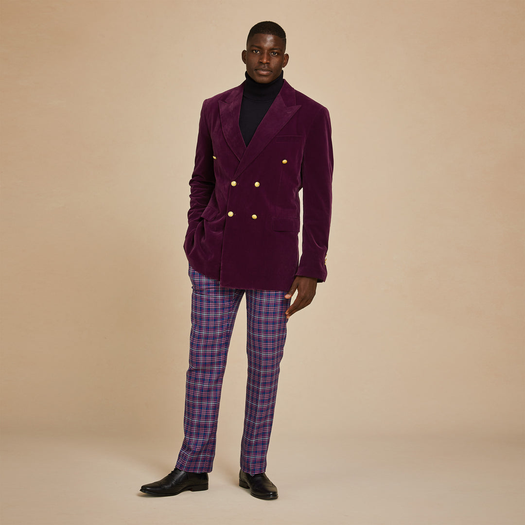 A man stands against a beige background wearing the Inserch DB Velvet Blazer BL502-166 in Deep Violet, paired with a black turtleneck, plaid trousers, and black dress shoes.