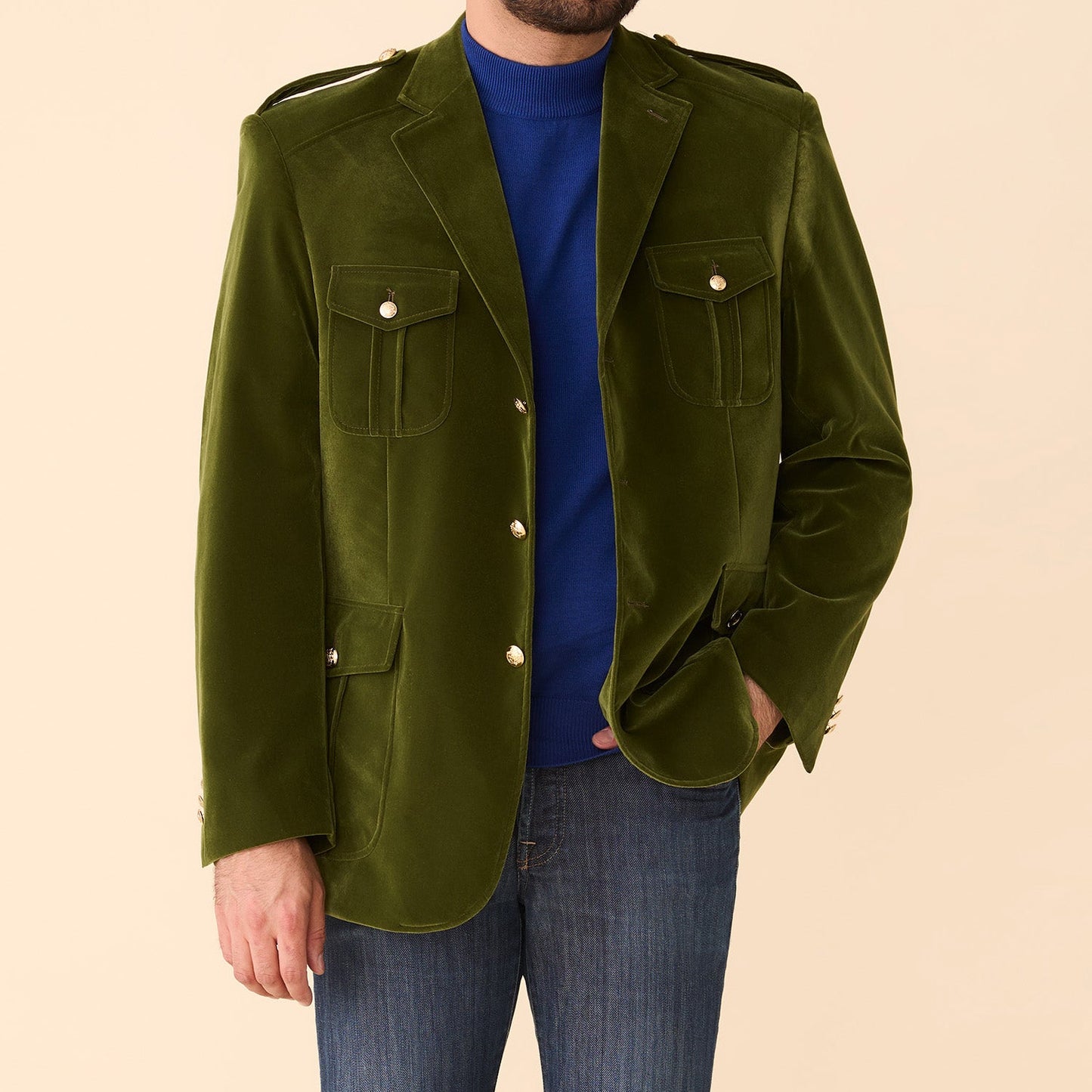 A man wears the Inserch Velvet Military Blazer BL569-19 Olive by Inserch over a blue turtleneck and blue jeans, standing against a beige background.