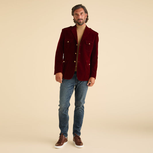 A man stands against a plain beige background wearing the Inserch Velvet Military Blazer BL569-31 in burgundy by Inserch, styled with a tan turtleneck sweater, blue jeans, and brown sneakers.