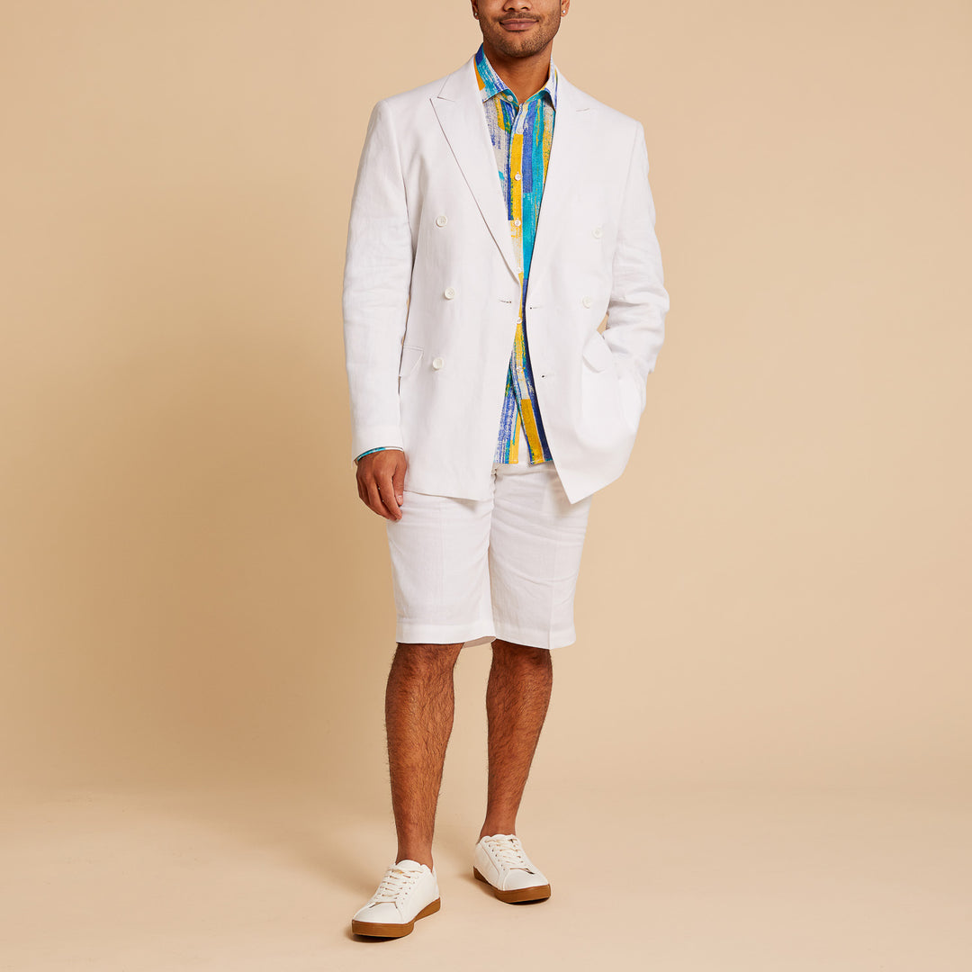 A man stands against a beige background wearing the Inserch Classic DB Linen Blazer BL660-02 White, paired with white shorts, a colorful shirt, and white sneakers with tan soles.