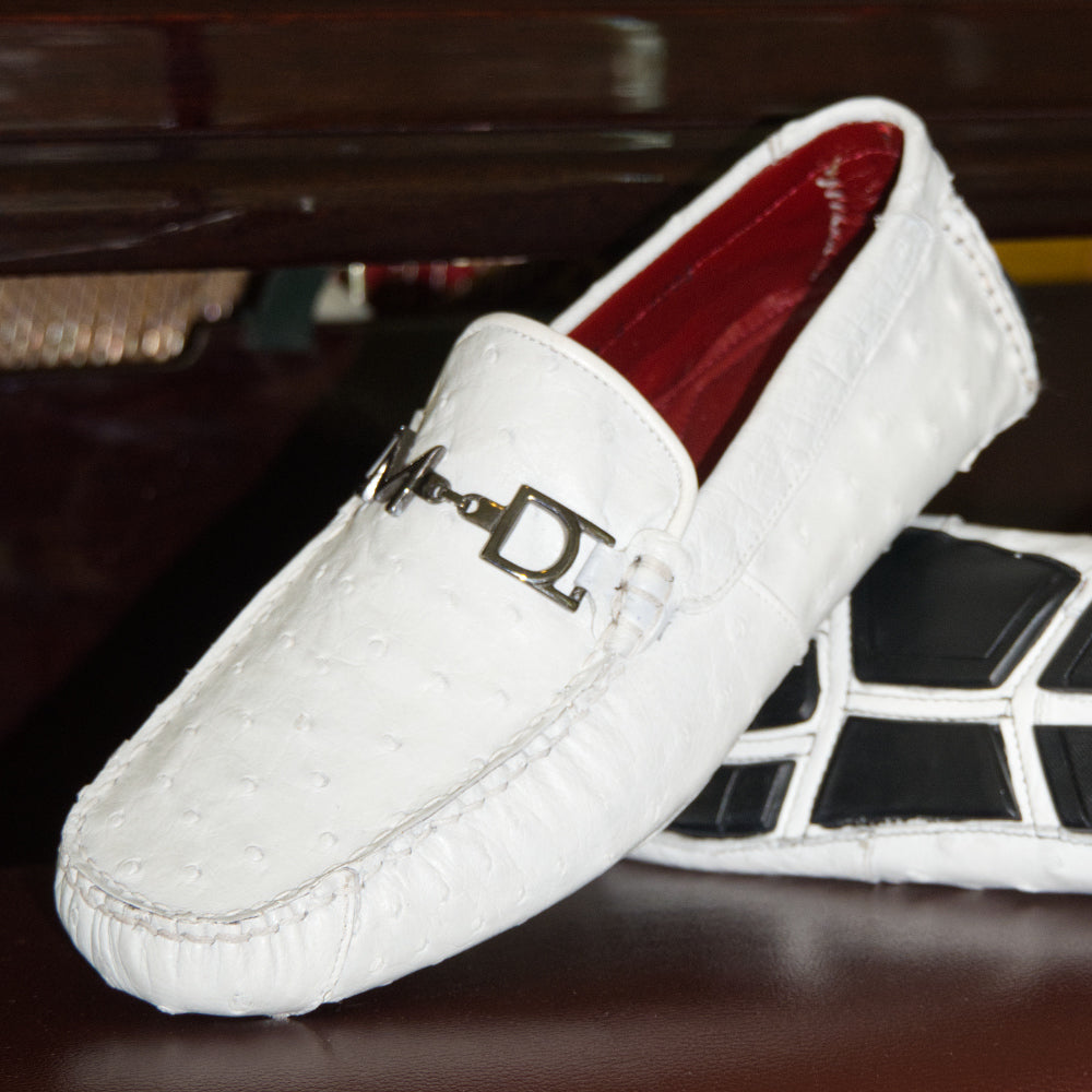 A pair of Bolonia White loafers by Marco Di Milano, featuring metal detailing on the front; one shoe stands upright while the other lies sideways to showcase the black and white sole—expertly crafted luxury moccasins designed to elevate any ensemble.