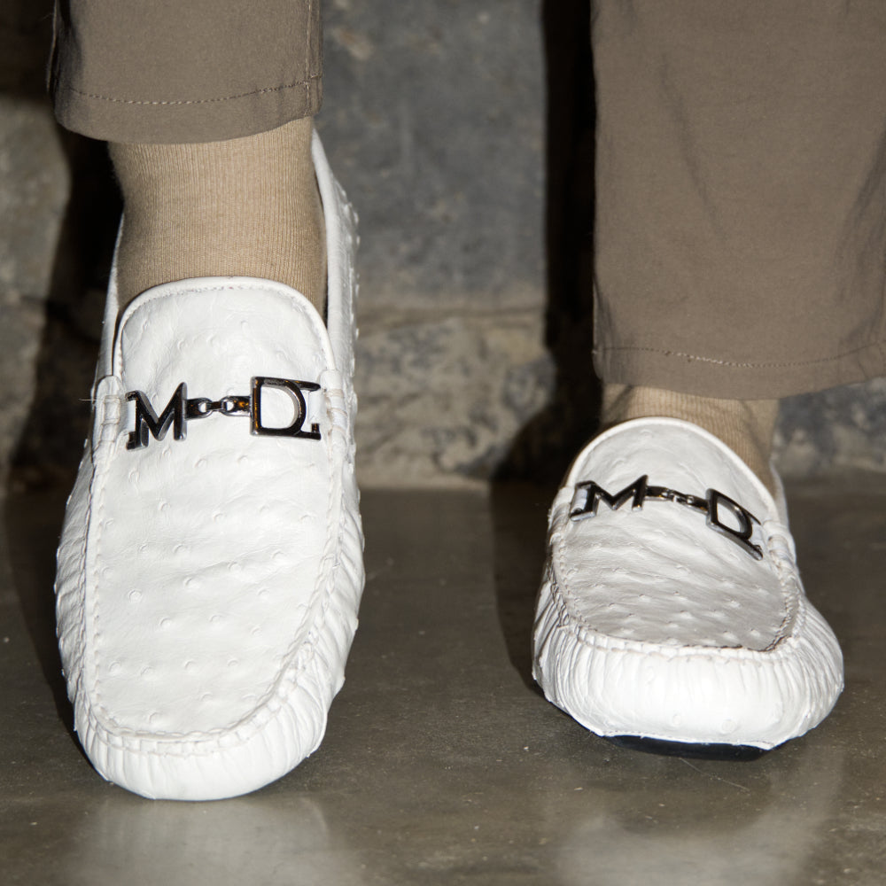 A pair of Bolonia White loafers by Marco Di Milano, featuring metal detailing on the front; one shoe stands upright while the other lies sideways to showcase the black and white sole—expertly crafted luxury moccasins designed to elevate any ensemble.