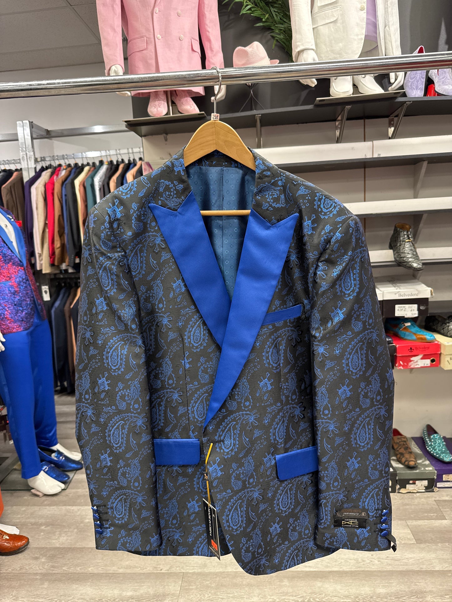 An EJ SAMUEL JACKET 27 CLEARANCE tuxedo jacket featuring a blue and black paisley pattern with blue lapels hangs on a wooden hanger in a clothing store; please note, this item is final sale with no exchanges or returns.